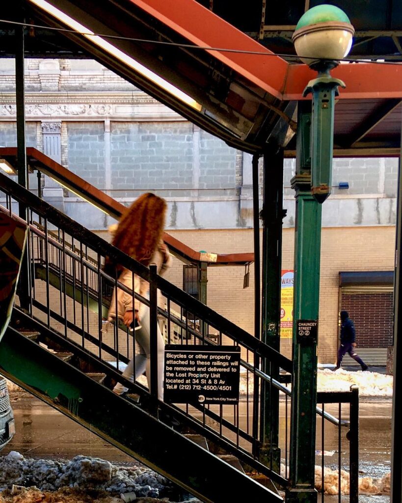 A woman walking down the staircase of the elevated subway at Chauncey Street on the J line on a winter day with dirty snow at the bottom