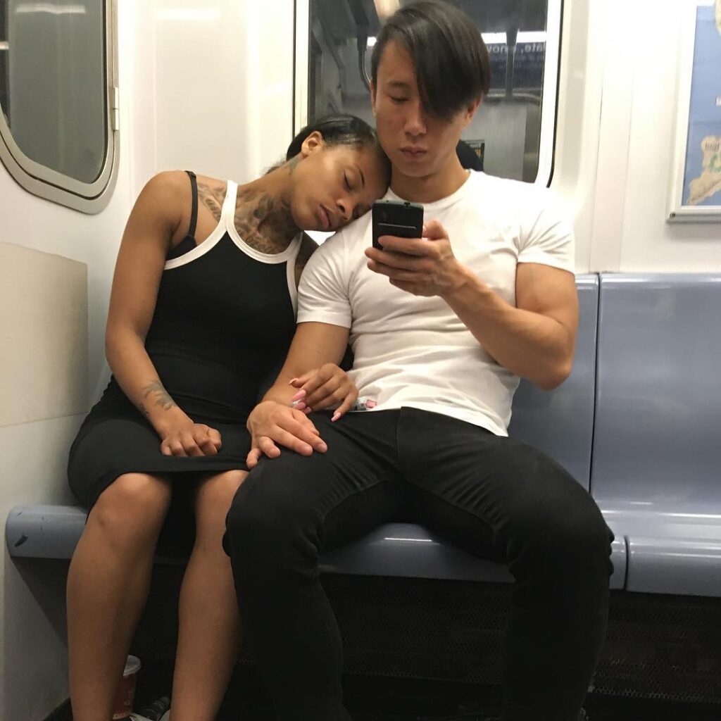 Black woman leaning on shoulder of white man in the corner of a subway car