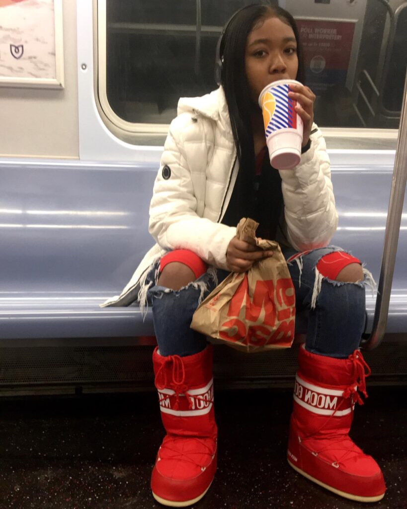 A woman with long braids sitting on a blue subway seat staring at the camera. She is drinking a soda and holding a McDonald's bag and wearing red boots