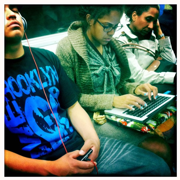 A photograph using a Hipstamatic filter of three people sitting on the subway, the middle one typing on a laptop.