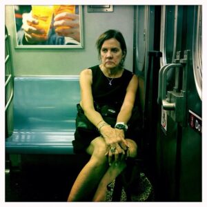 Photograph of a woman in a black dress sitting alone in the two-person seat on a subway car, an add for Budweiser in the top left shows two people sharing a toast